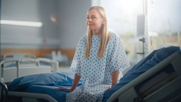 Modern Hospital Ward: Mooie blanke vrouwelijke patiënt rustend op een bed, volledig herstellend na succesvolle chirurgie, ziekte of Coronavirus glimlacht ze. Achter haar raam met een prachtig zonnig uitzicht — Stockfoto