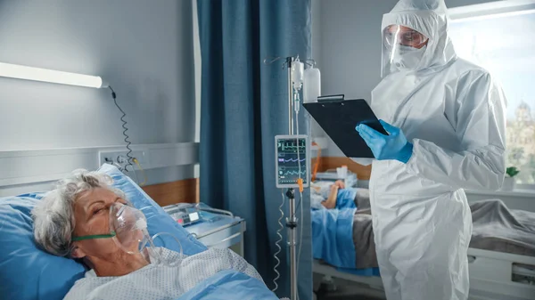 Ziekenhuis Coronavirus Emergency Department Ward: Dokter draagt Coverall opgeschreven vitale functies van een senior vrouwelijke patiënt met zuurstof masker rusten in bed. Medici, paramedici doen alles om levens te redden. — Stockfoto