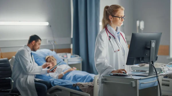 病棟:専門医または外科医は、医療用コンピュータを使用しています。背景にあるのは、ベッドで手術を成功させた後の回復患者のための近代的な機器クリニックの医師の座りとケア — ストック写真