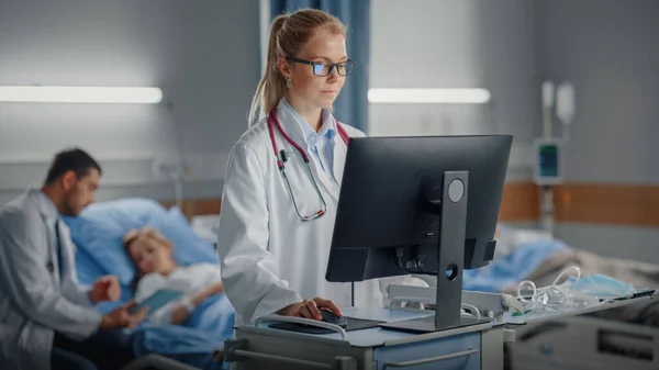 Krankenhausstation: Professionelle Ärztin oder Chirurgin verwendet medizinische Computer. Im Hintergrund sitzt ein Arzt, der sich nach erfolgreicher Operation im Bett erholt — Stockfoto