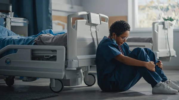 Coronavirus-Station auf der Intensivstation des Krankenhauses: Porträt einer traurigen, müden schwarzen Krankenschwester mit Gesichtsmaske, die auf einem Fußboden sitzt. Sorry für alle Patienten, die durch die Pandemie verloren wurden. Mutige Helden-Sanitäter retten Leben — Stockfoto