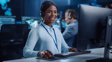 Neşeli Güzel Teknik Müşteri Destek Uzmanı, Meslektaşlar, Ekranlar ve Veri Sunucularıyla dolu Çağrı Merkezi Kontrol Odası 'nda bir bilgisayar üzerinde çalışırken Kulaklıkla Konuşuyor.