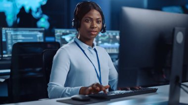 Neşeli Güzel Teknik Müşteri Destek Uzmanı, Meslektaşlar, Ekranlar ve Veri Sunucularıyla dolu Çağrı Merkezi Kontrol Odası 'nda bir bilgisayar üzerinde çalışırken Kulaklıkla Konuşuyor.