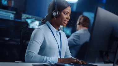 Neşeli Güzel Teknik Müşteri Destek Uzmanı, Meslektaşlar, Ekranlar ve Veri Sunucularıyla dolu Çağrı Merkezi Kontrol Odası 'nda bir bilgisayar üzerinde çalışırken Kulaklıkla Konuşuyor.