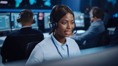 Neşeli Güzel Teknik Müşteri Destek Uzmanı, Meslektaşlar, Ekranlar ve Veri Sunucularıyla dolu Çağrı Merkezi Kontrol Odası 'nda bir bilgisayar üzerinde çalışırken Kulaklıkla Konuşuyor.