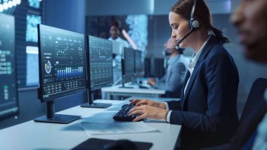 Mutlu Güzel Teknik Müşteri Destek Uzmanı, İş Arkadaşları ve Ekranlarla Dolu Karanlık İzleme ve Kontrol Odasında Bilgisayar üzerinde Çalışırken Kulaklık Çağrısı Yapıyor.