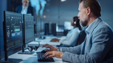 Masasında birden fazla bilgisayar görüntüsüyle oturan Ciddi Profesyonel Teknik Denetçi Portresi. Arka planda, meslektaşları Sistem Kontrol ve İzleme Merkezi 'nde çalışıyor..