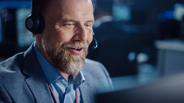 Bearded Happy Senior Technical Support Specialist spricht auf einem Headset, während er an einem Computer in einem dunklen Überwachungs- und Kontrollraum mit Computerbildschirmen und Datenservern arbeitet. — Stockfoto