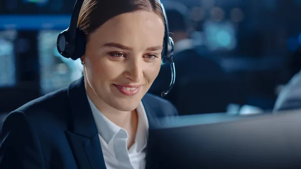 Primer plano Retrato de un feliz y hermoso especialista en soporte técnico al cliente que habla con auriculares mientras trabaja en una computadora en una oscura sala de monitoreo llena de colegas y pantallas. —  Fotos de Stock