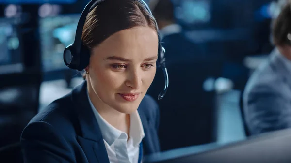 Happy Confident Technical Customer Support Specialist Having a Headset Call while Working on a Computer in a Dark Monitoring and Control Room Filled with Colleagues and Display Screens. — Zdjęcie stockowe