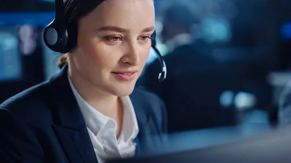 Närbild Porträtt av en glad vacker teknisk kundsupport specialist talar på ett headset medan du arbetar på en dator i ett mörkt övervakningsrum fylld med kollegor och bildskärmar. — Stockfoto