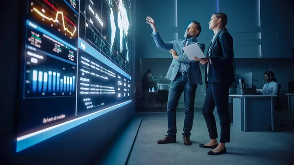 Projectmanager en Computer Science Engineer praten tijdens het gebruik van Big Screen Display en een laptop, Toont Infrastructuur Infografische Gegevens. Controle- en monitoringkamer voor telecommunicatiebedrijven. — Stockfoto