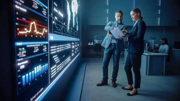 Project Manager och Computer Science Engineer Talk while Using Big Screen Display och en bärbar dator, visar infrastruktur Infographics Data. Telekommunikationsföretagets kontroll- och övervakningsrum. — Stockfoto