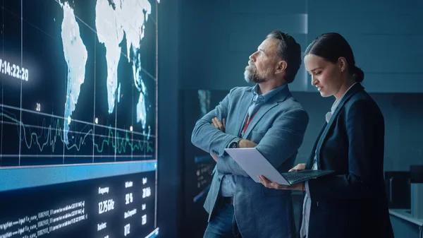 Project Manager och Computer Science Engineer Talk while Using Big Screen Display och en bärbar dator, visar infrastruktur Infographics Data. Telekommunikationsföretagets kontroll- och övervakningsrum. — Stockfoto