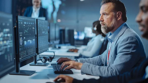 Retrato del controlador técnico profesional serio sentado en su escritorio con múltiples pantallas de computadora ante él. En segundo plano Sus colegas que trabajan en el Centro de Control y Monitoreo de Sistemas. —  Fotos de Stock