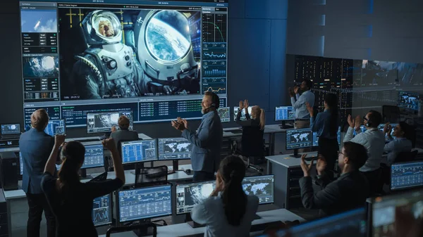 Groupe de personnes au centre de contrôle de mission Établir une connexion vidéo réussie sur un grand écran avec un astronaute à bord d'une station spatiale. Les scientifiques des commandes de vol se lèvent et frappent des mains. — Photo