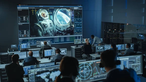 Groep mensen in Mission Control Center Vestig succesvolle videoverbinding op een groot scherm met een astronaut aan boord van een ruimtestation. Flight Control Wetenschappers zitten voor Computers. — Stockfoto