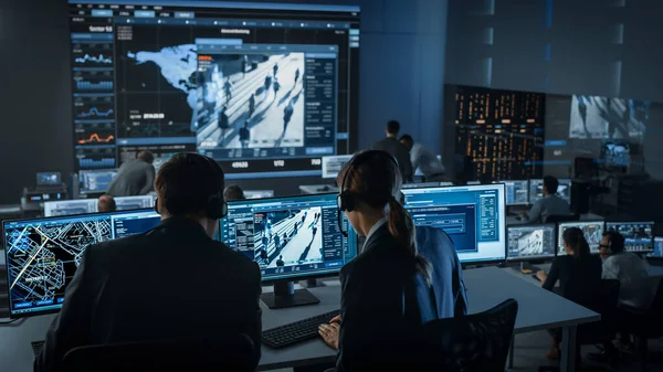女警官在一个大型数字屏幕上有全球地图追踪的警察监测中心的一台装有闭路电视录像录像的计算机上工作。员工坐在"大数据"展示前 — 图库照片