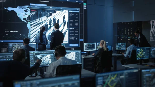 Middelbare mannelijke en vrouwelijke overheidsfunctionarissen bespreken werkzaken naast een groot digitaal scherm met satellietbewakingsbeelden met gezichtsherkenningssoftware. Politie Onderzoekers aan het werk. — Stockfoto