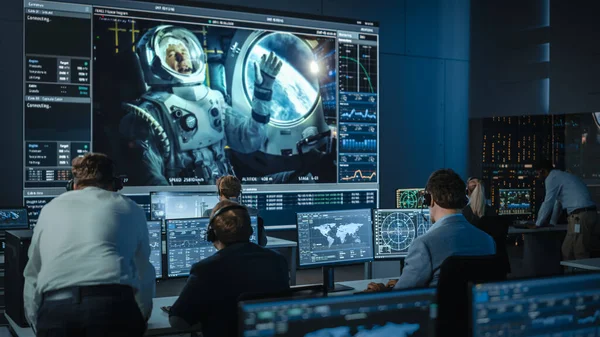 Grupo de Pessoas no Centro de Controle de Missão Estabeleça uma Conexão de Vídeo de Sucesso em uma Tela Grande com um Astronauta a bordo de uma Estação Espacial. Cientistas de Controle de Voo sentam-se na frente dos computadores. — Fotografia de Stock