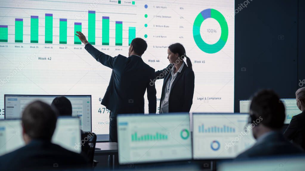 Two Traders Having a Meeting in a Modern Monitoring Office with Analytics Feed on a Big Digital Screen. Monitoring Room with Brokers and Finance Specialists Sit in Front of Computers. Colleagues Talk.