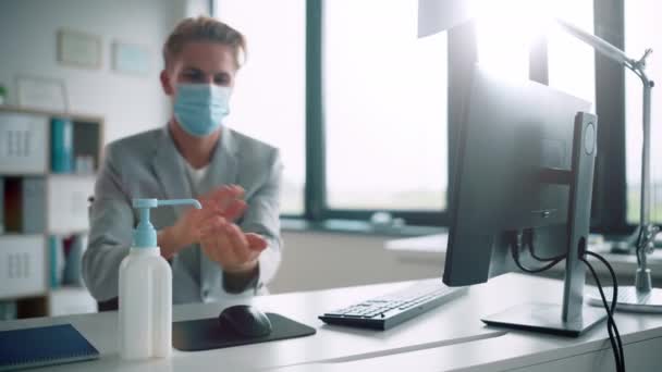 Молодой человек в маске для лица использует средство для дезинфекции рук в офисе — стоковое видео