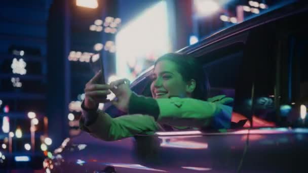 興奮した女性は夜の車の後部座席から窓の外を見ている 働くネオンサインで街の通りがいかに美しいかを驚きのスマートフォンで写真やビデオを撮る女性 — ストック動画