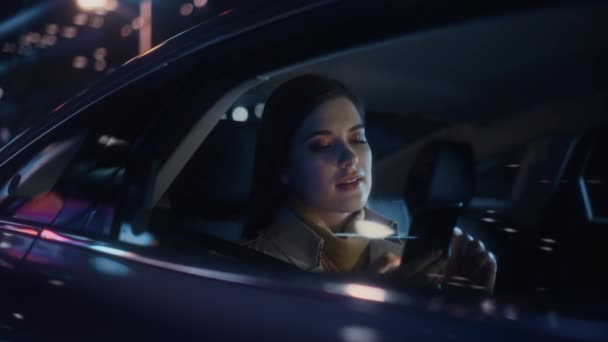 スタイリッシュな女性は夜にタクシーの後部座席で家に通勤しています 働くネオンサインで都会の街の車の中でスマートフォンを使って窓から外を見る美しい女性乗客 — ストック動画