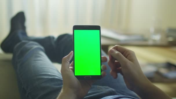 Людина лежить на дивані вдома і використовує Android телефон з зеленим екраном в портретному режимі на лапі . — стокове відео