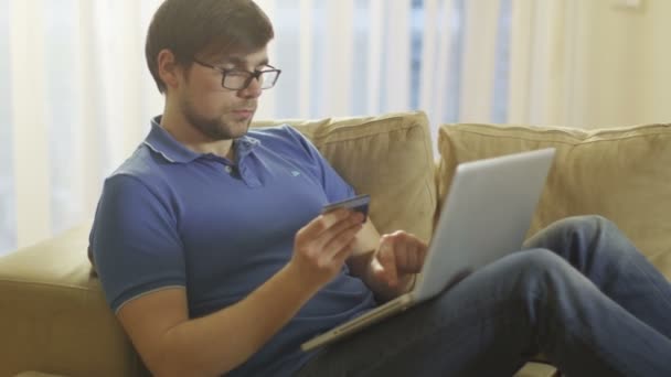 El hombre acostado en el sofá y el uso de ordenador portátil para compras en línea en el hogar — Vídeo de stock
