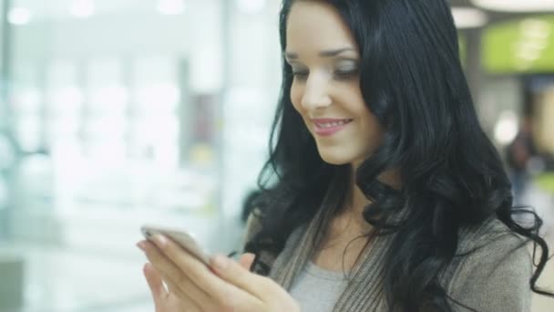 Menina morena nova com belo sorriso perfeito está usando o telefone móvel no shopping. Tiro de mão . — Vídeo de Stock