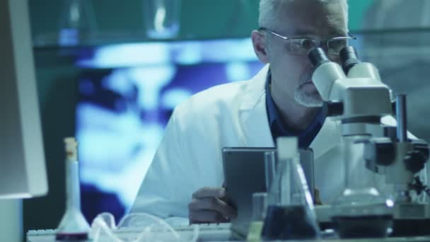 Scientifique regarde à travers le microscope et l'utilisation de la tablette en laboratoire — Video