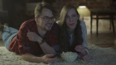 Young Happy Smiling Couple are Laying on the Floor and Watching TV and Eating Popcorn.