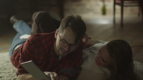 Young Happy Smiling Couple estão deitados no chão e usando Tablet PC para entretenimento em casa no horário da noite. Estilo de vida casual . — Vídeo de Stock