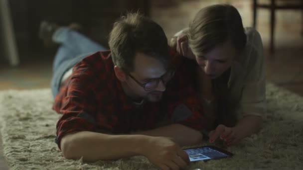 Young Happy Smiling Couple estão deitados no chão e usando Tablet PC para entretenimento em casa no horário da noite. Estilo de vida casual . — Vídeo de Stock