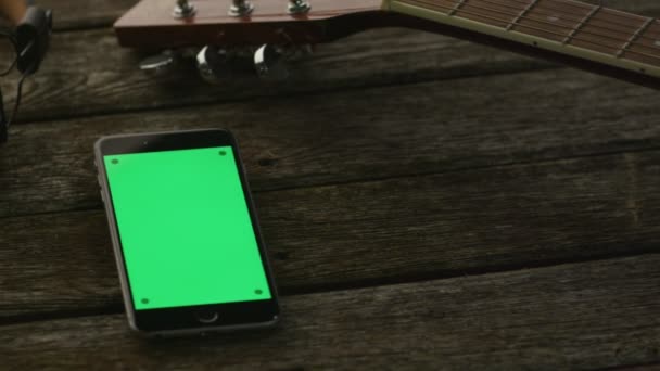 Телефон с зеленым экраном в портретном режиме, лежащий на деревянном столе рядом с гитарой и наушниками. Цезарь Ливни — стоковое видео