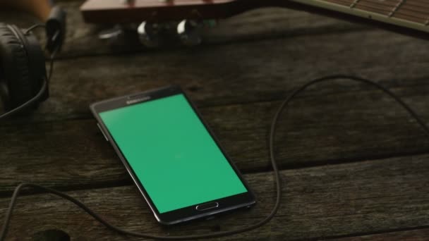 Android-телефон с зеленым экраном в портрете, лежащий на столе Wooden рядом с гитарой и наушниками. Цезарь Ливни — стоковое видео