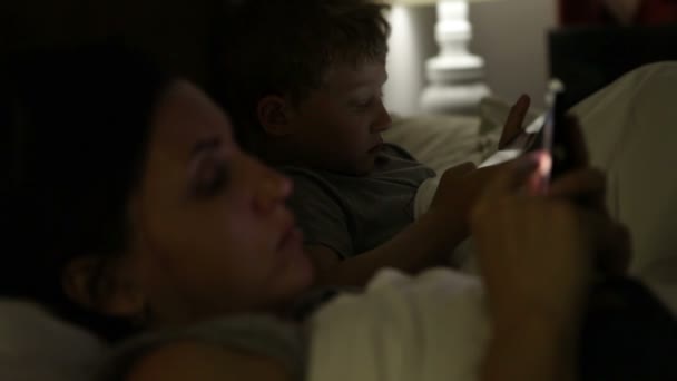 Мать с сыном, лежа в постели и посмотрите в их электронных устройств — стоковое видео