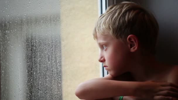 Ensam liten pojke ser regndroppar genom fönsterglas — Stockvideo