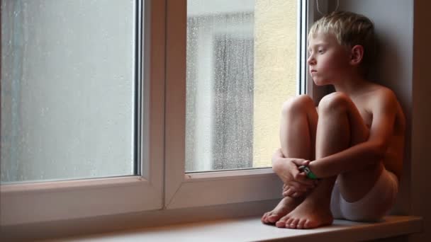 Alone little boy looks raindrops through window glass — Stock Video
