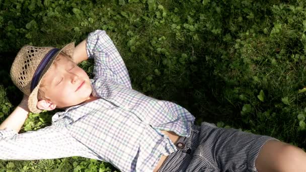 Avslappende gutt under det grønne treet som fanger de varme solstrålene som skinner gjennom bladene – stockvideo