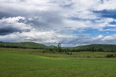 Önünde boş bir tarla ile tepelerin ardında dramatik bir gökyüzü görülebilir.