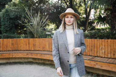  Gri ceketli, şapkalı, kazaklı, kot pantolonlu sarışın bir kadın parkta yürüyor. Ahşap bankların olduğu modern bir parkta yürümek. Hafta sonu kaçamağı.