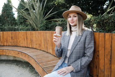  Avrupalı görünümlü bir kadın uzun tahta bir bankta oturur ve kahve içer. Şapkalı, ceketli, kazaklı ve kotlu bir kız cam bardaktan su içer.