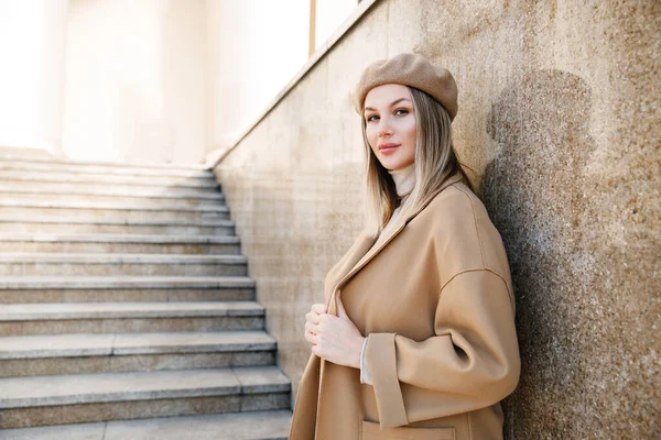 Young beautiful sweet woman in coat, beret, sweater, jeans, light shoes. Posing on the stairs near the theater building. The blonde girl is alone in the street.