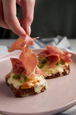 Kadınların elleri Parma jambonunu tost veya brusket peyniri, kabak ve domatesle süslüyor. Et ve tahıl ekmeğiyle yararlı yiyecekler. Pembe tabakta sağlıklı yemek..