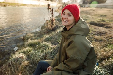 Genç bir turist kız yağmurlukla, şapkayla ve turist botlarıyla kameraya gülüyor. Güneşli hava nehrinin kenarında çimlerin üzerinde oturuyor. Çevresini çevreleyen doğayı düşünerek. 