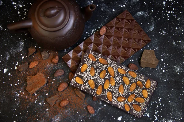 Yanında kahverengi çaydanlık, kakao parçaları, kakao çekirdekleri ve dökülmüş dökülen siyah ve beyaz bir zemin olan badem ve susam tohumlu iki çikolatalı süt.