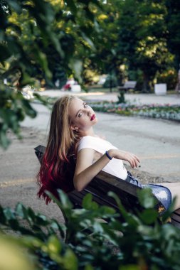 Young girl sits on a bench in a summer green garden. A girl of European appearance, blond with blue eyes.