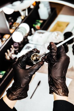 Siyah eldivenli ustanın elleri makyaj sıvısı ve fırçayla dolu bir kap tutuyor..